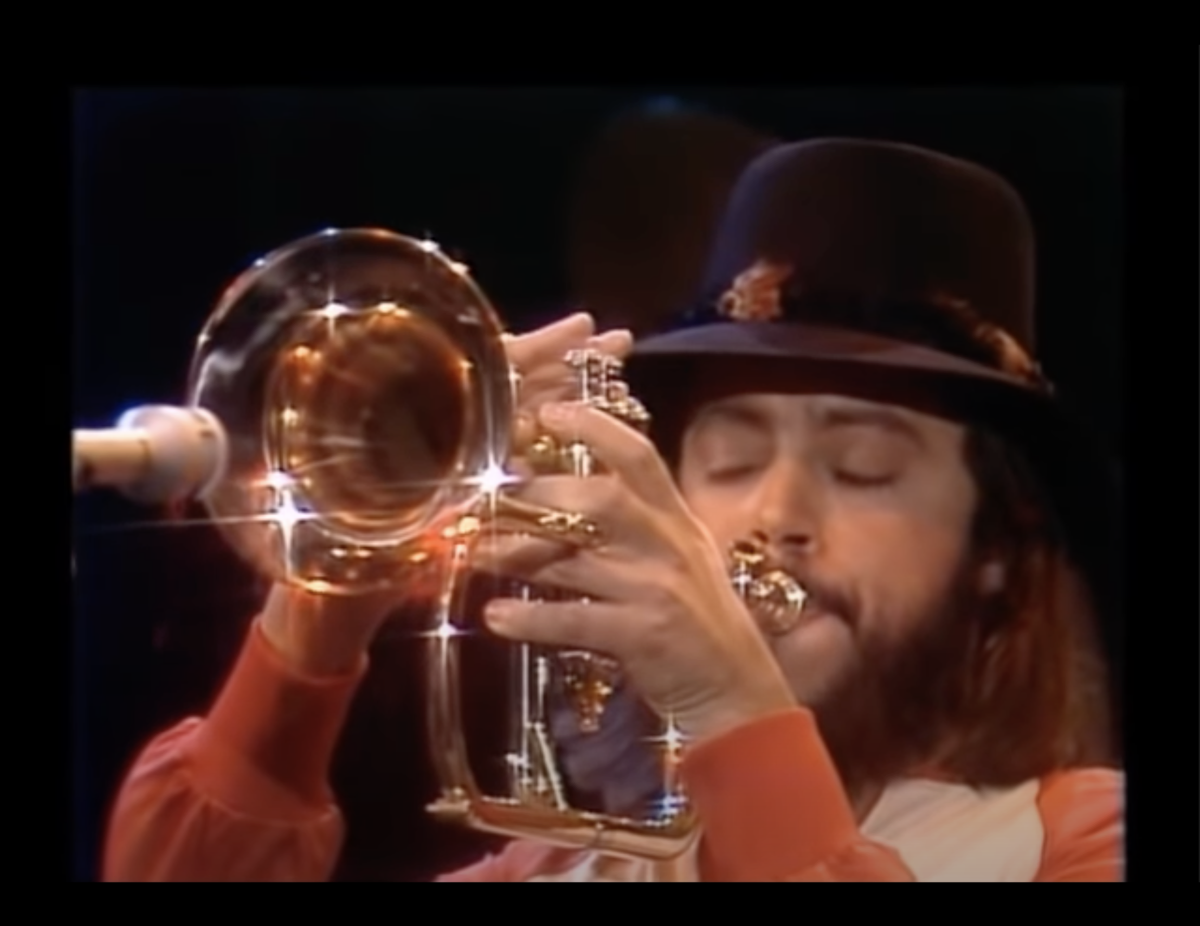 Photo: still from a video. A very 1970s looking setup. A man in a hat plays a brass instrument with a small band in a TV studio.